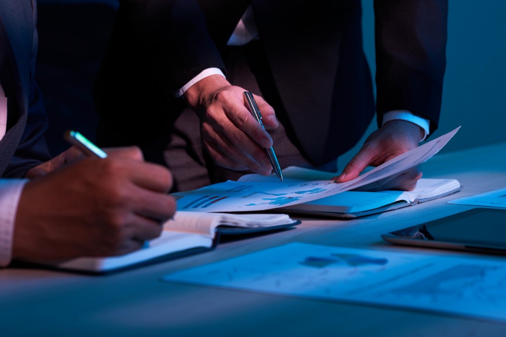 Pessoas em reunião analisando e assinando documentos, com caneta e mãos sobre papéis abertos sobre uma mesa, em ambiente corporativo com iluminação azul.