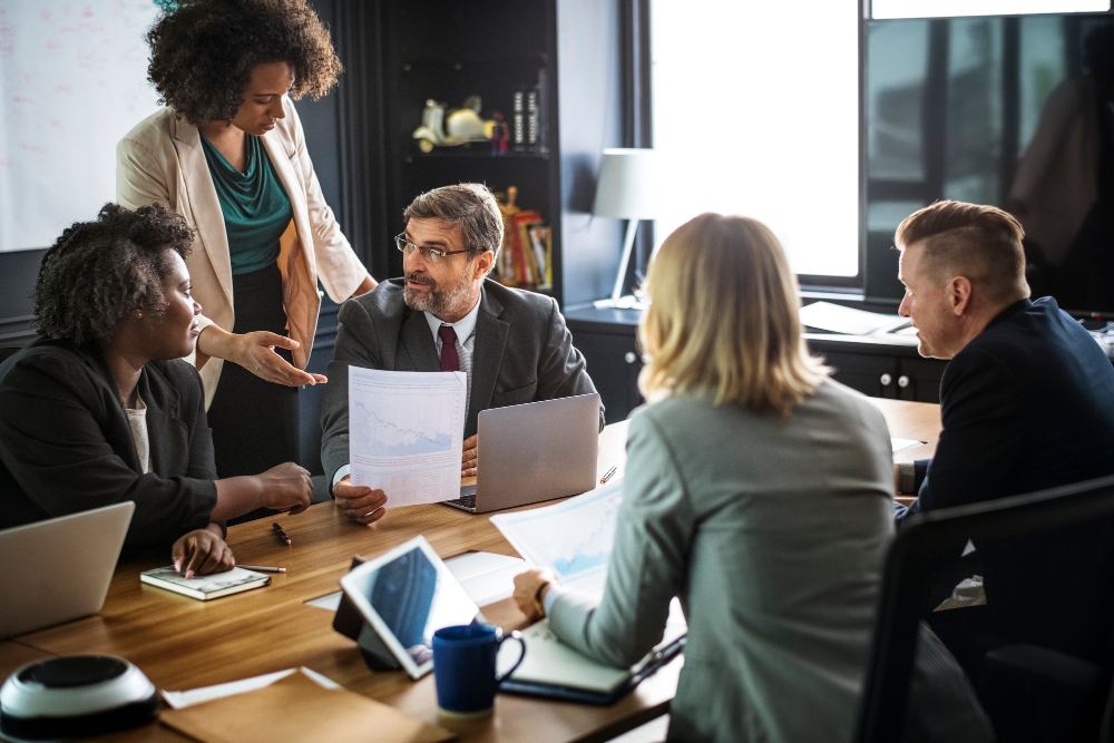 Reunião de equipe em escritório: pessoas discutem ideias ao redor de uma mesa com laptops, papéis e um mapa, enquanto uma mulher em pé aponta informações para o grupo. Ambiente corporativo.