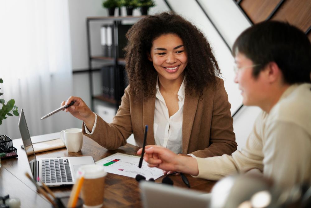 Reunião de trabalho em escritório moderno: uma mulher com cabelo cacheado sorri enquanto explica um plano, com canetas e papéis sobre a mesa, ao lado de dois colegas
