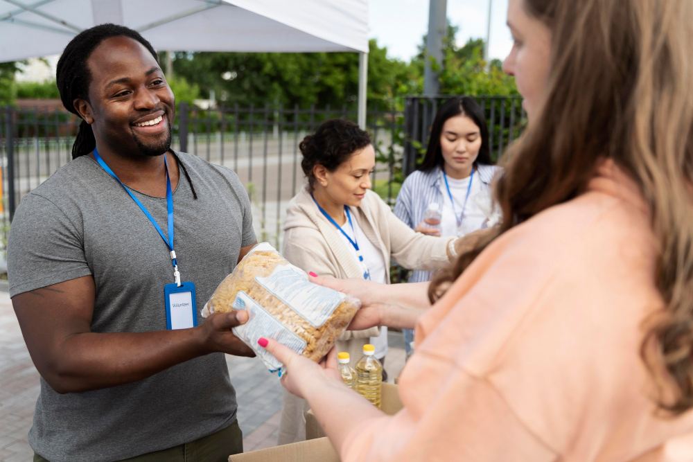 Voluntários em um evento ao ar livre embaixo de uma tenda, entregando alimentos e apoiando a distribuição de doações, com pessoas sorrindo e conversando.