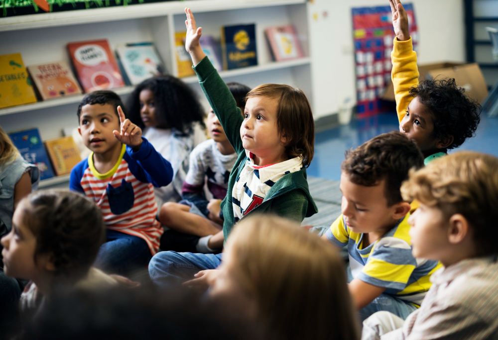 Crianças em sala de aula, sentadas em círculo, participam de atividade educativa e levantam as mãos para responder durante a aula