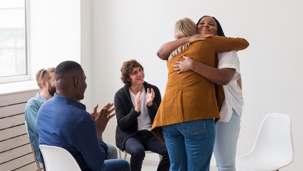 Equipe em reunião se abraçando em clima de apoio e união, com pessoas sentadas e duas pessoas de pé trocando um abraço no escritório, promovendo bem-estar no trabalho.