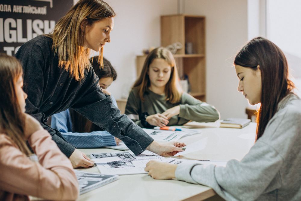 Professoras e estudantes em sala de aula trabalhando em atividades impressas, discutindo e realizando exercícios sobre materiais gráficos em uma mesa durante o estudo