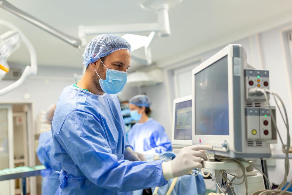 Médico cirurgião em sala de cirurgia com roupa estéril, máscara e luvas, operando um equipamento de monitoramento em um hospital moderno com equipe ao fundo.