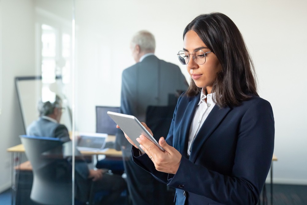 Mulher executiva com óculos e terno azul usando um tablet em ambiente corporativo moderno, enquanto outras pessoas trabalham ao fundo em escritórios