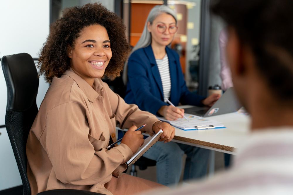 Mulher em primeiro plano sorrindo em uma reunião de trabalho, escrevendo em um tablet e usando caneta, enquanto duas pessoas ao fundo analisam documentos em uma sala de escritório