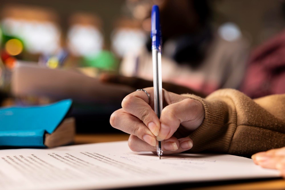 Pessoa segurando uma caneta e escrevendo em uma folha de papel sobre uma mesa, com objetos e material escolar ao fundo, em ambiente de estudo ou escritório.