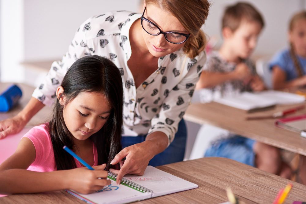 Professora orienta uma criança em sala de aula, enquanto ela desenha e escreve em um caderno com lápis de cor, ao fundo outros alunos e materiais escolares.