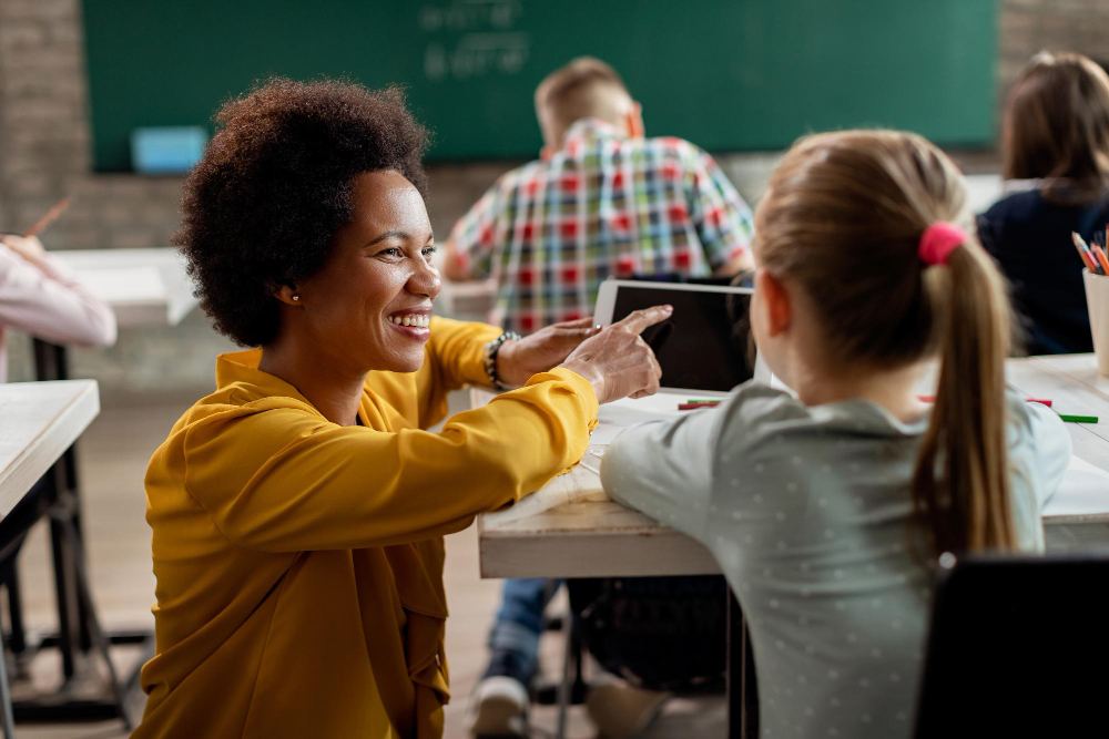 Professora sorridente interagindo com estudantes em sala de aula, com alunos sentados em carteiras e quadro verde ao fundo, durante uma atividade pedagógica.