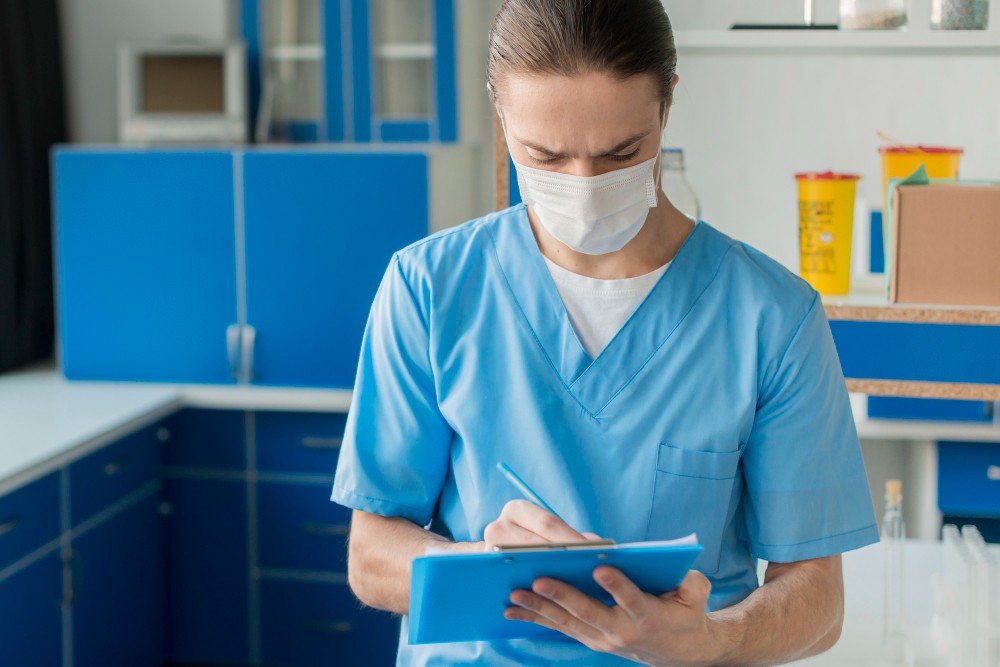 Profissional de saúde usando máscara cirúrgica em clínica, segurando prancheta azul e escrevendo durante atendimento, com ambiente hospitalar ao fundo.