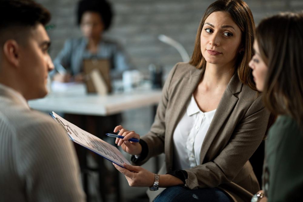 Mulher em reunião de trabalho, segurando documentos e apresentando um plano enquanto conversa com colegas em ambiente de escritório, com foco na abordagem profissional
