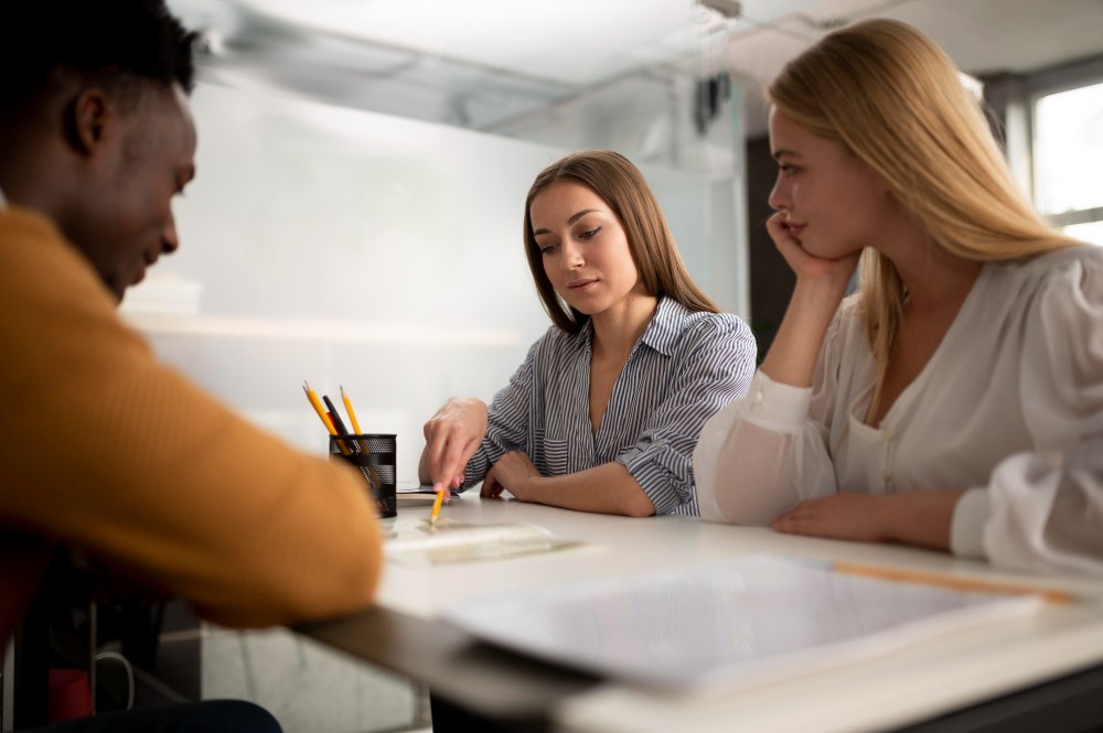 Reunião em escritório com três pessoas discutindo um projeto sobre uma mesa, com papéis e canetas enquanto avaliam ideias